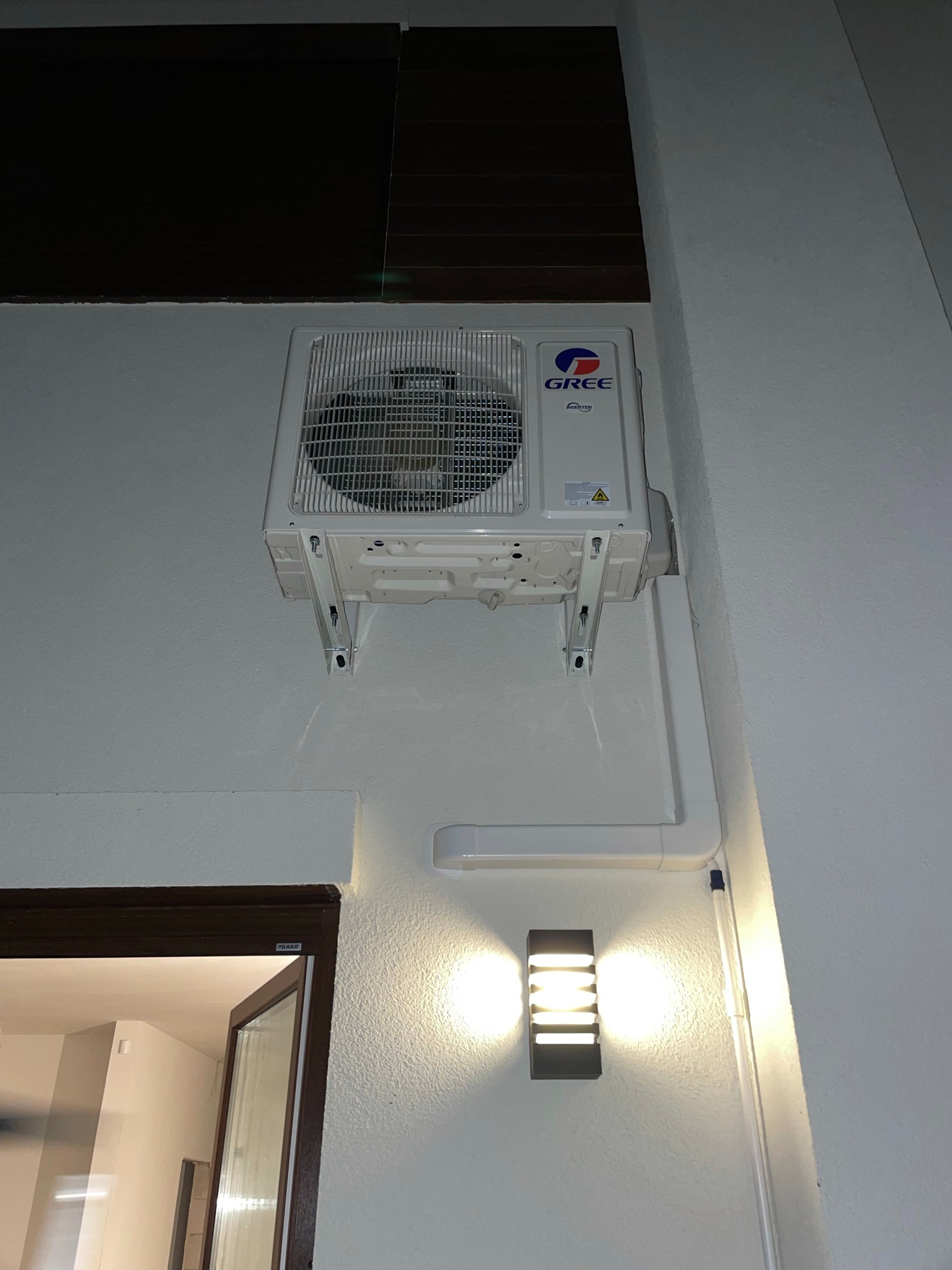 Zamontowana klimatyzacja marki Gree na białej ścianie z widocznymi rurami odprowadzającymi oraz kinkietem oświetlającym.