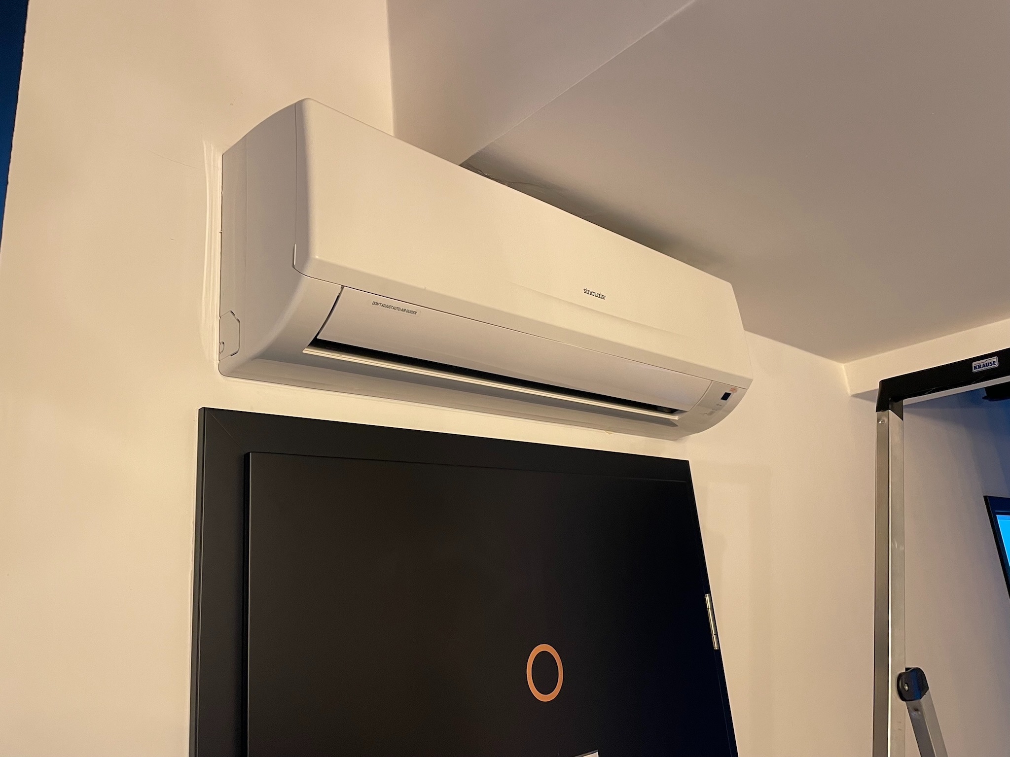 Biały klimatyzator Sinclair zamontowany na ścianie nad czarnymi drzwiami z okrągłym uchwytem, widoczny fragment drabiny w tle.