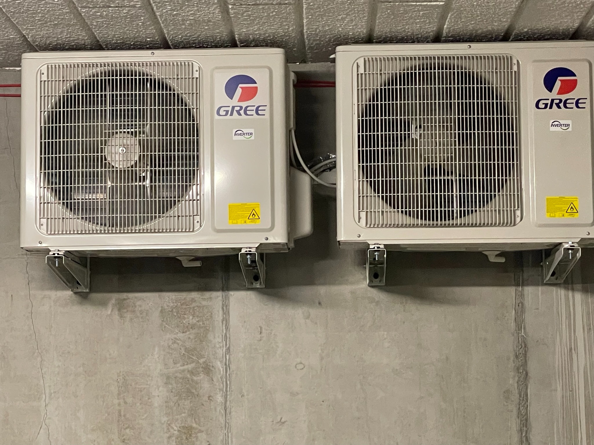 Dwa identyczne klimatyzatory GREE zamontowane na szarej, surowej ścianie betonowej, widoczne mocowania i przewody.