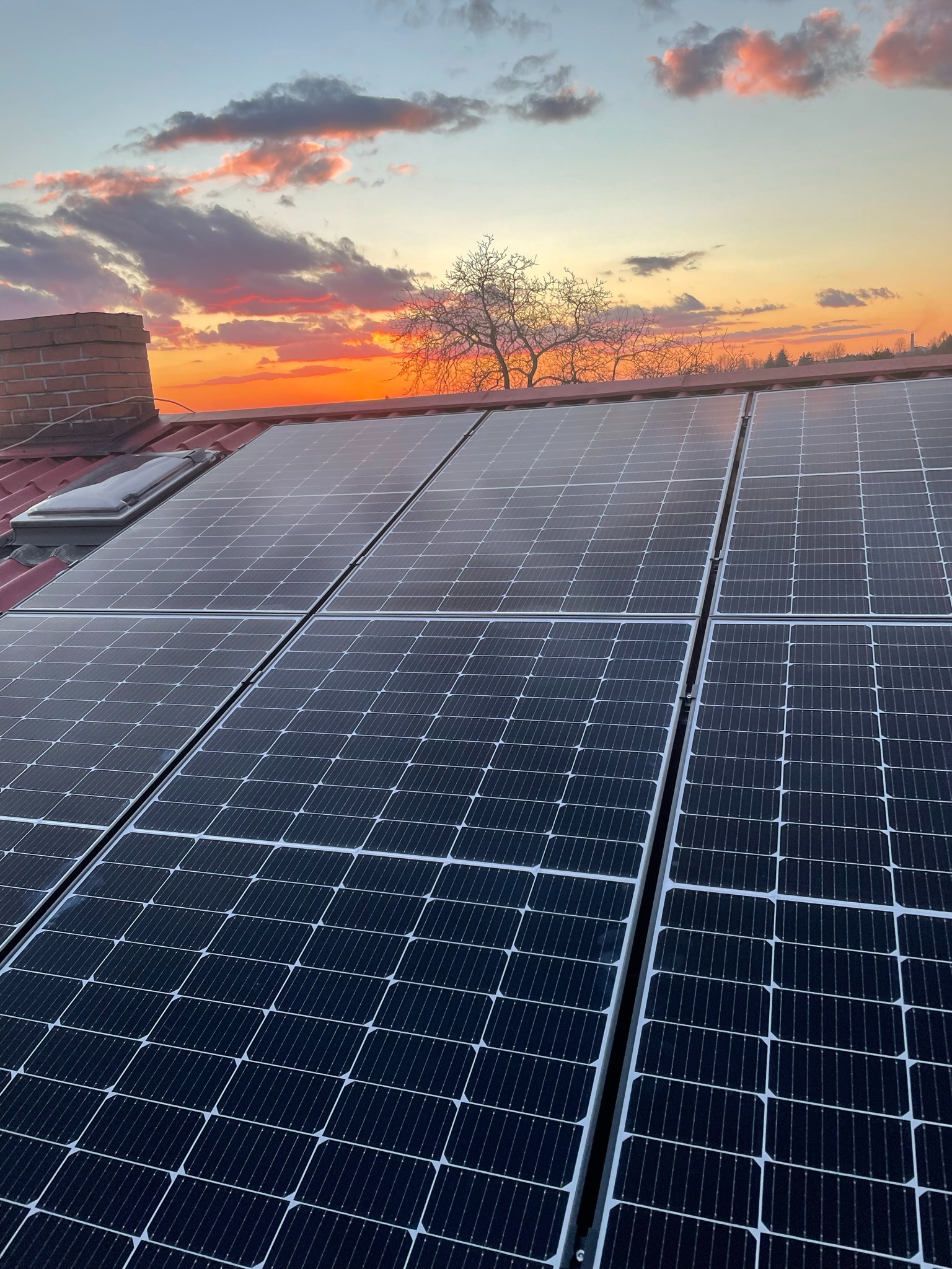 Panele fotowoltaiczne na dachu domu, widoczne przy zachodzącym słońcu, z kominem i oknem dachowym.