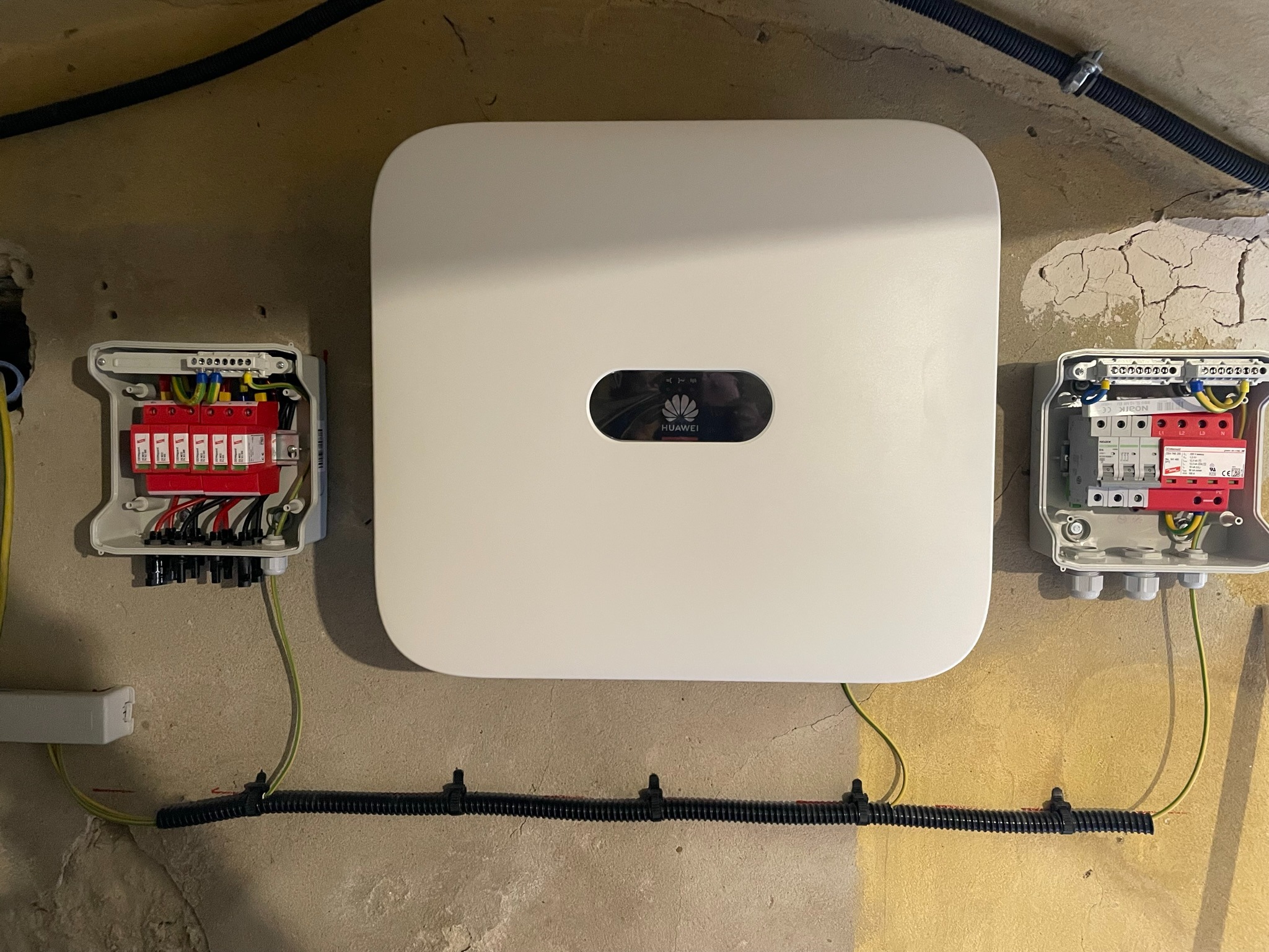 Biała obudowa falownika Huawei zamontowana na ścianie z widocznymi zabezpieczeniami przeciwprzepięciowymi w szarych skrzynkach po bokach i przewodami ochronnymi uziemiającymi.