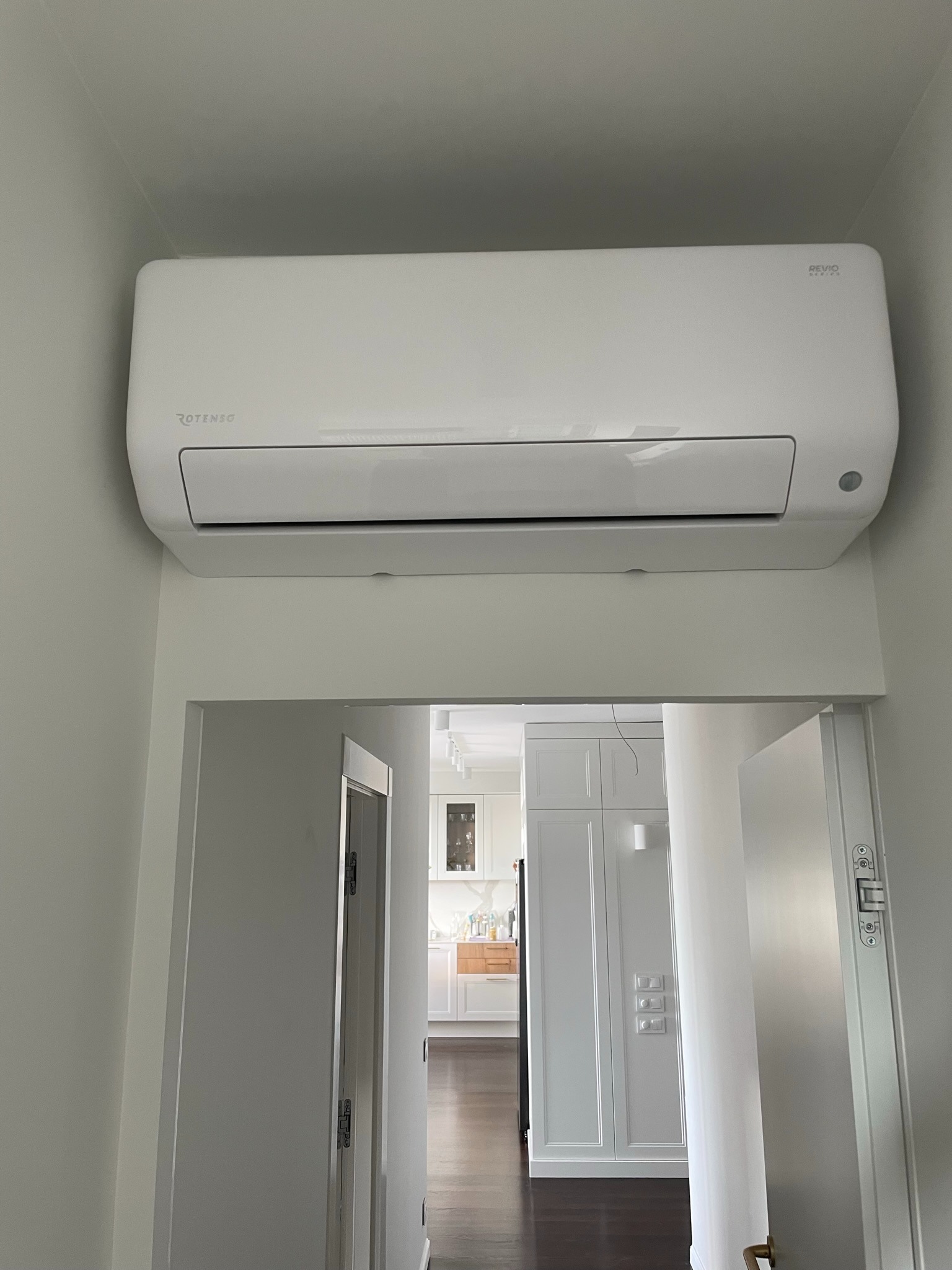 Zamontowany biały klimatyzator marki Rotenso w mieszkaniu w Będzinie, widok z dołu na urządzenie zawieszone nad otwartymi drzwiami do jasnej kuchni.