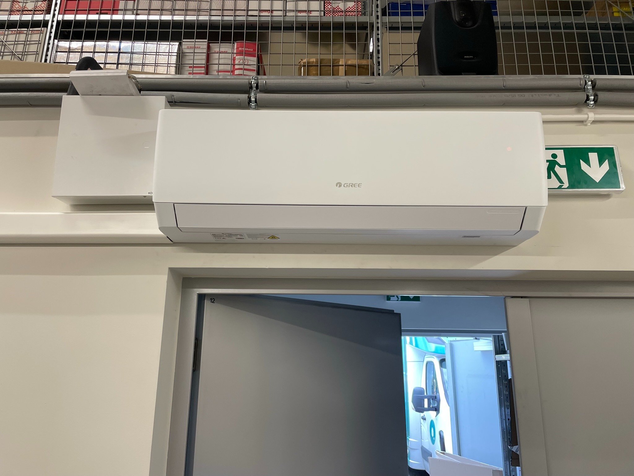 Biały klimatyzator marki Gree zamontowany na ścianie w pomieszczeniu magazynowym, widoczny znak ewakuacyjny i regały z towarem.