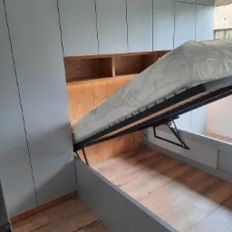 Szare szafy na wymiar z drewnianymi półkami i łóżkiem z podnoszonym stelażem, materac w folii ochronnej, widoczny mechanizm podnoszenia.