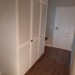 Biała szafa wnękowa z lamelkowymi drzwiami na brązowej podłodze, widoczna biała ściana i drzwi.