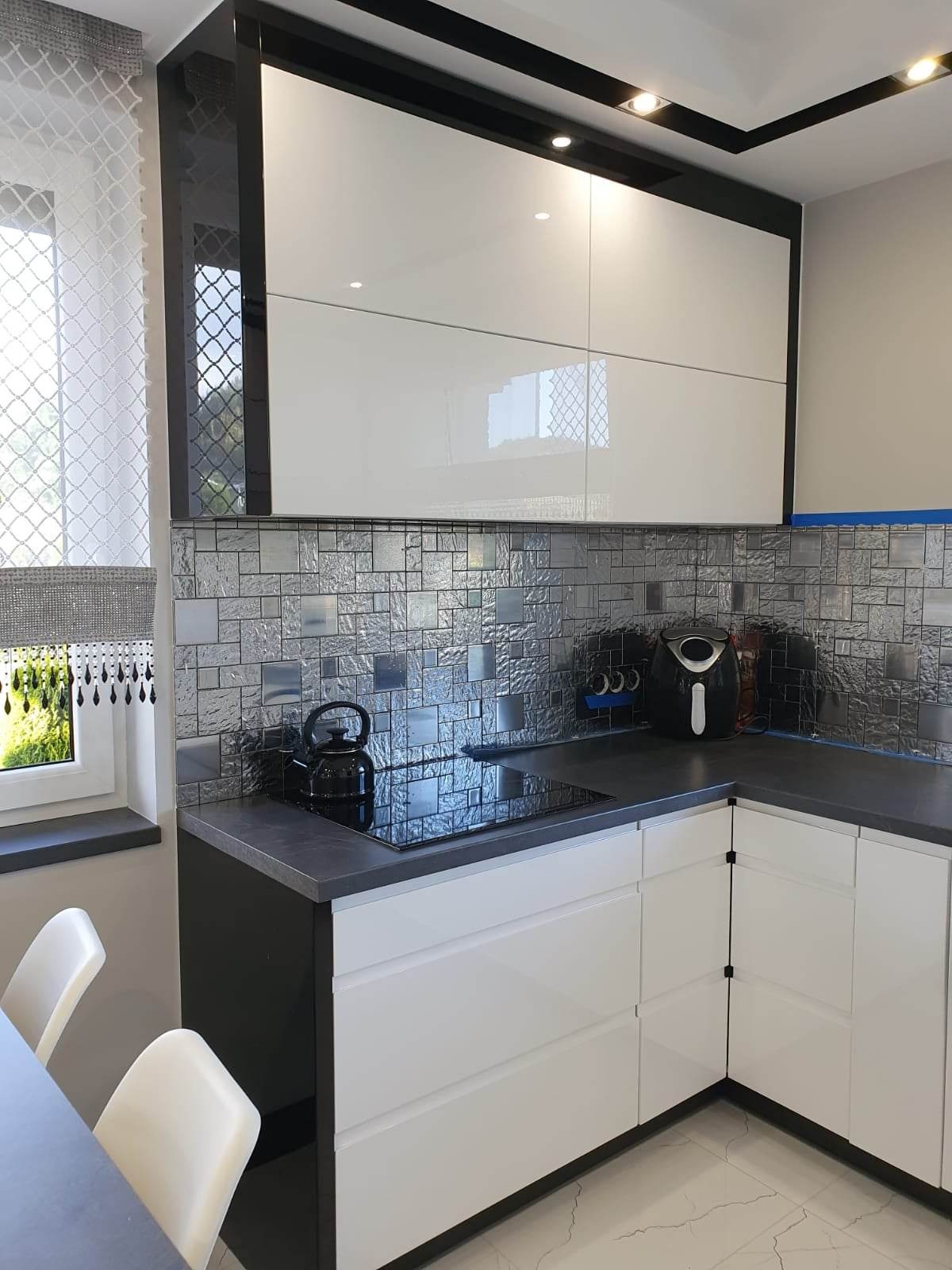 Nowoczesna kuchnia: białe, lakierowane szafki wiszące z czarnymi obramowaniami, czarny blat, srebrna mozaika na ścianie, indukcyjna płyta grzewcza, czajnik i frytkownica beztłuszczowa na blacie...