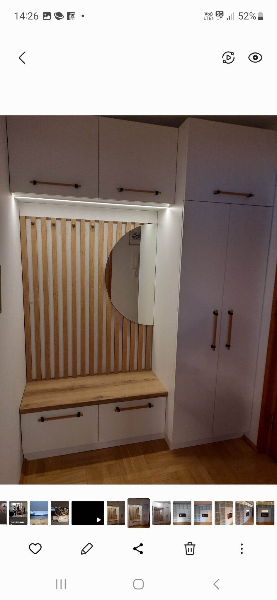 Biała szafa wnękowa z drewnianymi akcentami, siedziskiem i okrągłym lustrem w przedpokoju, oświetlenie LED podkreśla pionowe drewniane listwy na ścianie.