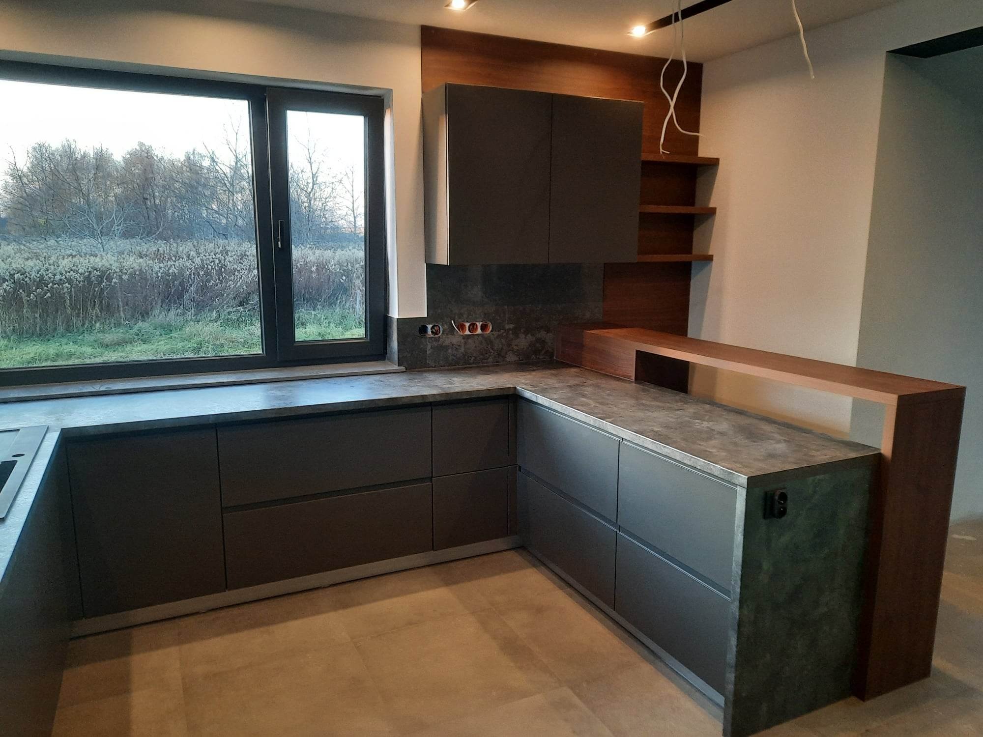 Nowoczesna, szara zabudowa kuchenna z blatem o fakturze betonu, widok na krajobraz za oknem, częściowo wykończone drewniane elementy i widoczne przewody elektryczne.