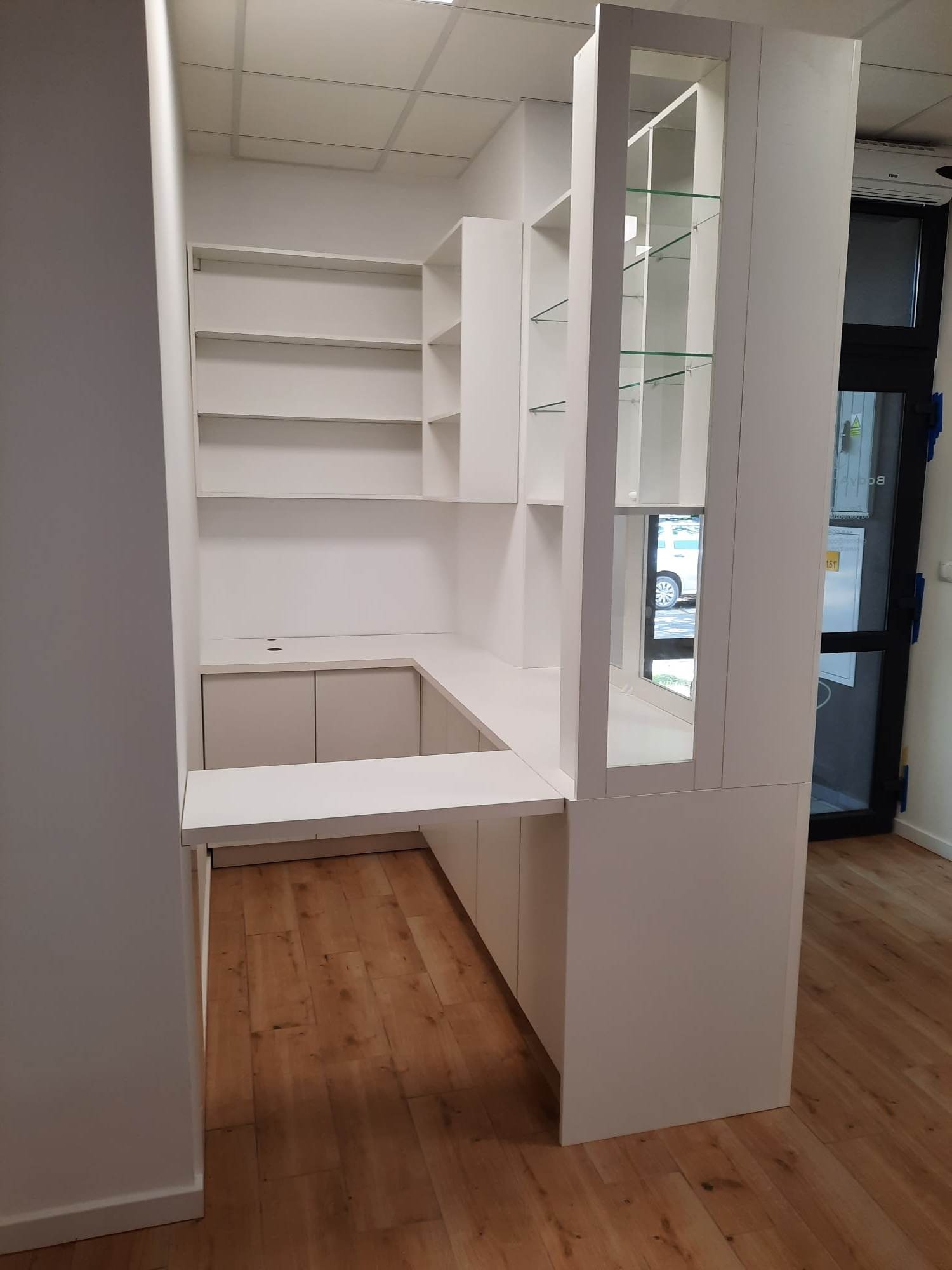 Biała zabudowa meblowa z półkami i przeszkloną witryną, drewniana podłoga, widok z boku na fragment pomieszczenia.