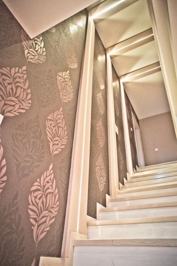 Klatka schodowa z drewnianymi stopniami, oświetleniem LED i tapetą w liście, sfotografowana z dołu, ukazująca perspektywę w górę.