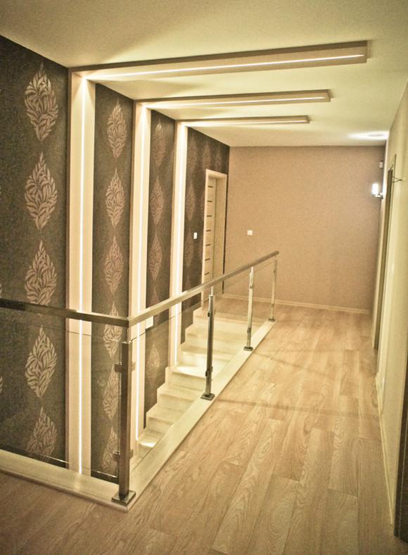 Klatka schodowa z drewnianymi schodami i szklanymi balustradami, oświetlona paskami LED na ścianach z wzorzystą tapetą, drewniana podłoga i oświetlenie sufitowe.