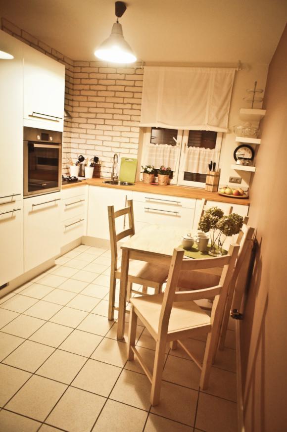 Jasna kuchnia z białymi szafkami, drewnianym blatem, ceglaną ścianą i stołem z krzesłami. Na blacie kuchennym widoczne akcesoria i kwiaty w doniczkach.