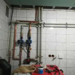 Instalacja hydrauliczna z miedzianymi i plastikowymi rurami, dwoma niebieskimi pompami obiegowymi, widoczne narzędzia i farba na czerwonym stole roboczym, białe płytki na ścianie.