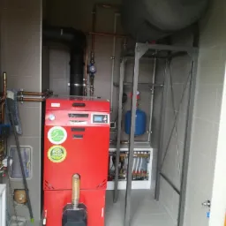 Czerwony piec na paliwo stałe z naklejką 'eco design', podłączony do instalacji w kotłowni z szarymi płytkami, widoczny zbiornik akumulacyjny i osprzęt instalacyjny.