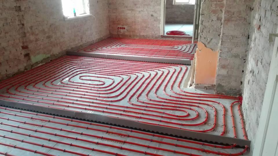 Czerwone rury ogrzewania podłogowego ułożone na szarym podłożu w surowym wnętrzu z ceglanymi ścianami i widocznymi instalacjami elektrycznymi.