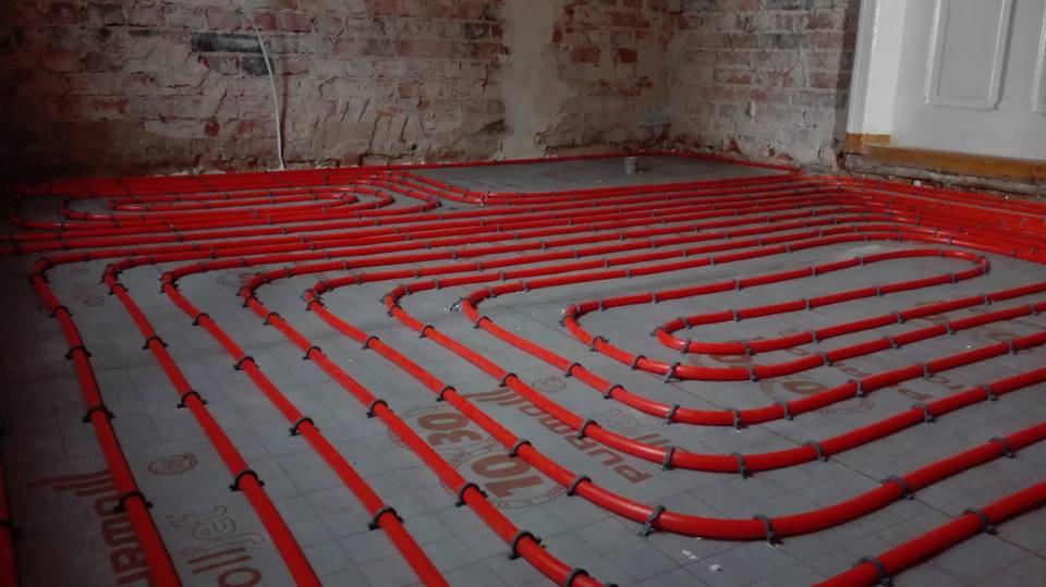 Czerwone rury ogrzewania podłogowego ułożone na szarych płytach izolacyjnych z widocznymi napisami, na tle ściany z surowej cegły i białych drzwi.