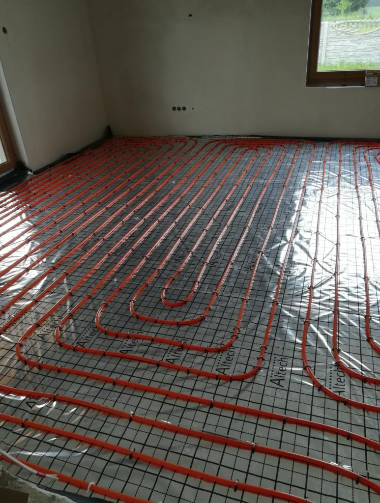 Pomarańczowe rury ogrzewania podłogowego ułożone na folii z nadrukowaną siatką, widoczne w surowym wnętrzu z oknem.