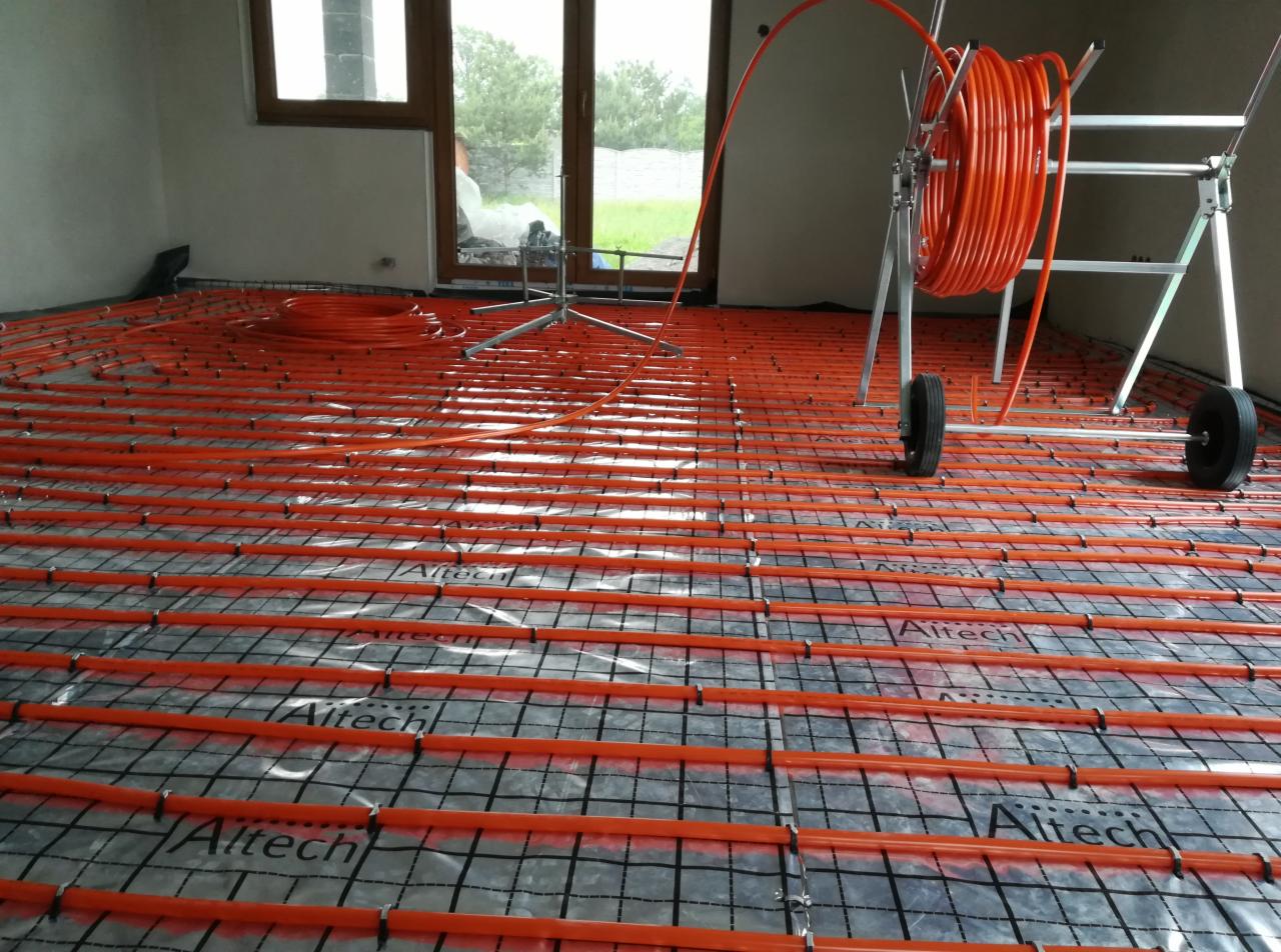 Pomarańczowe rury ogrzewania podłogowego rozłożone na czarnej siatce z folią, widoczny zwijak do rur i okno z widokiem na zewnątrz.