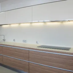 Nowoczesna kuchnia: dolne szafki z drewnopodobnym fornirem i metalowymi listwami, górne szafki białe, podświetlenie LED pod szafkami, minimalistyczny zlew i płyta indukcyjna.