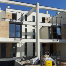 Nowoczesny budynek wielorodzinny w trakcie wykańczania elewacji, z widocznymi elementami dekoracyjnymi z imitacji kamienia, stolarką okienną w kolorze ciemnego brązu i przygotowanym materiałem...