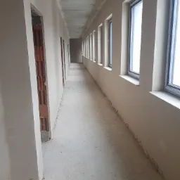 Długi korytarz w stanie surowym, ściany pokryte świeżym tynkiem, widoczne okna i otwory drzwiowe z ceglaną konstrukcją, betonowy sufit, podłoga z resztkami zaprawy.