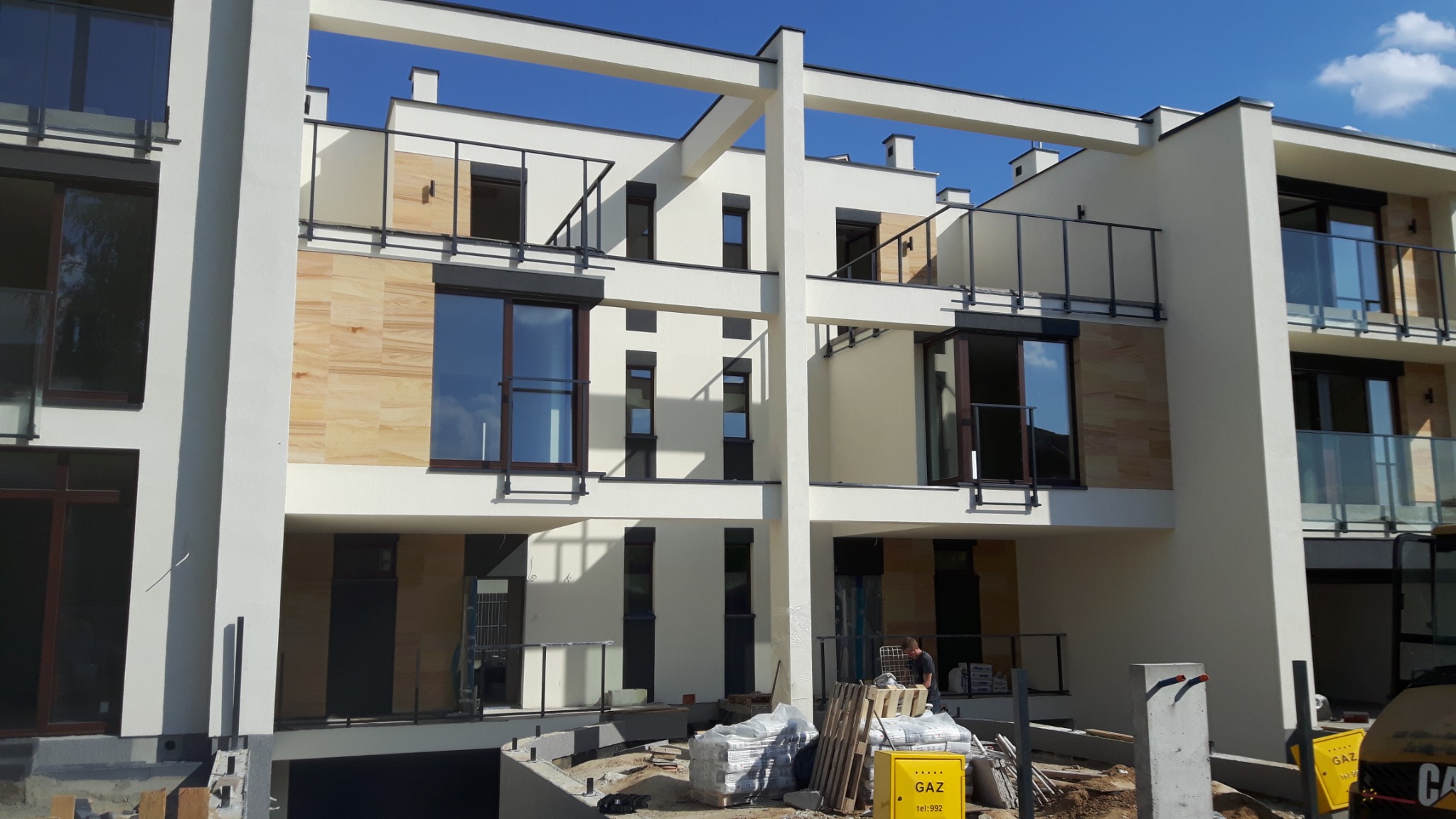 Nowoczesny budynek wielorodzinny w trakcie wykańczania elewacji, z widocznymi elementami dekoracyjnymi z imitacji kamienia, stolarką okienną w kolorze ciemnego brązu i przygotowanym materiałem...