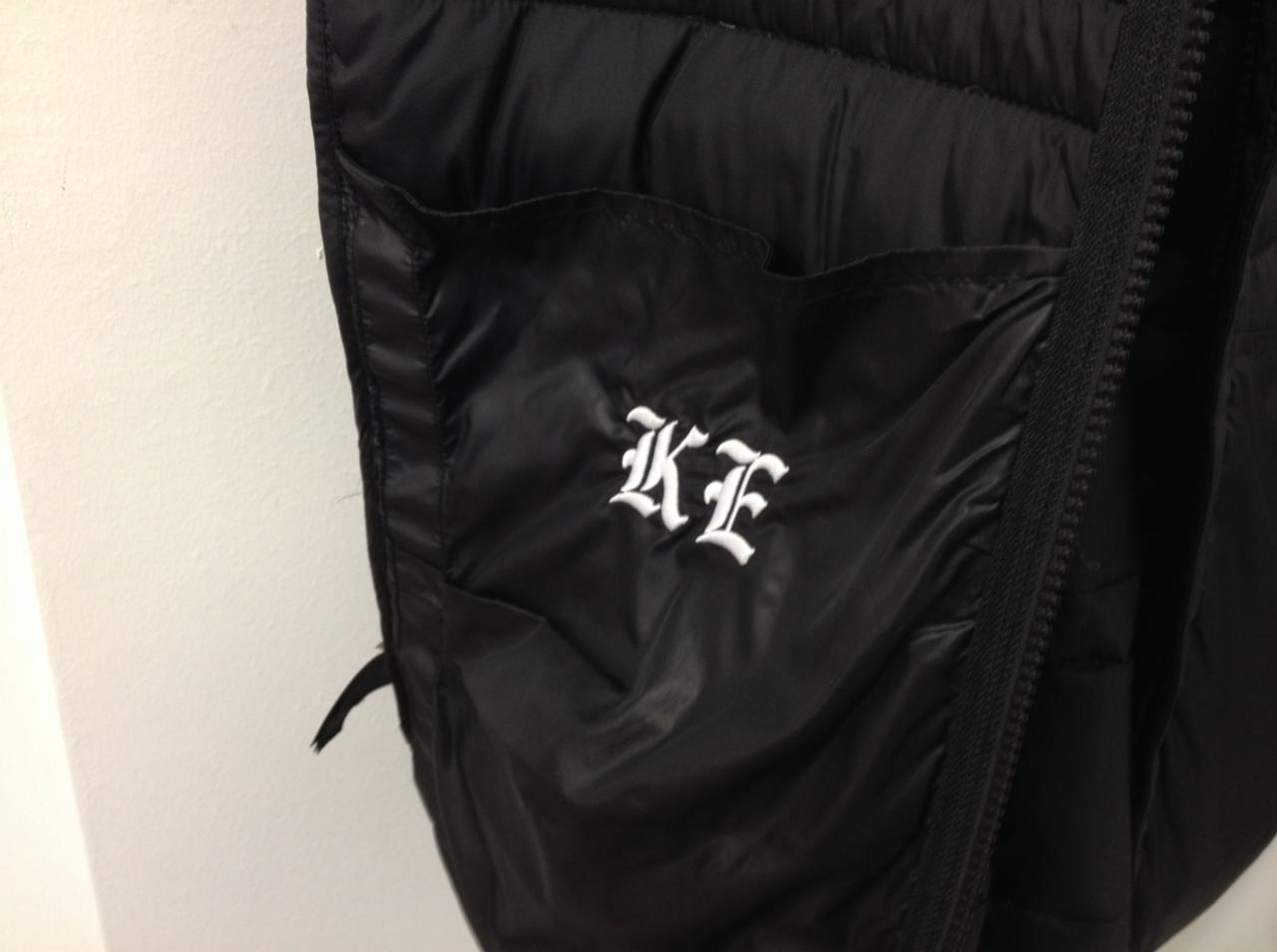 Czarna, błyszcząca kamizelka z kieszenią, na której wyhaftowane są białe, ozdobne inicjały 'KE'.