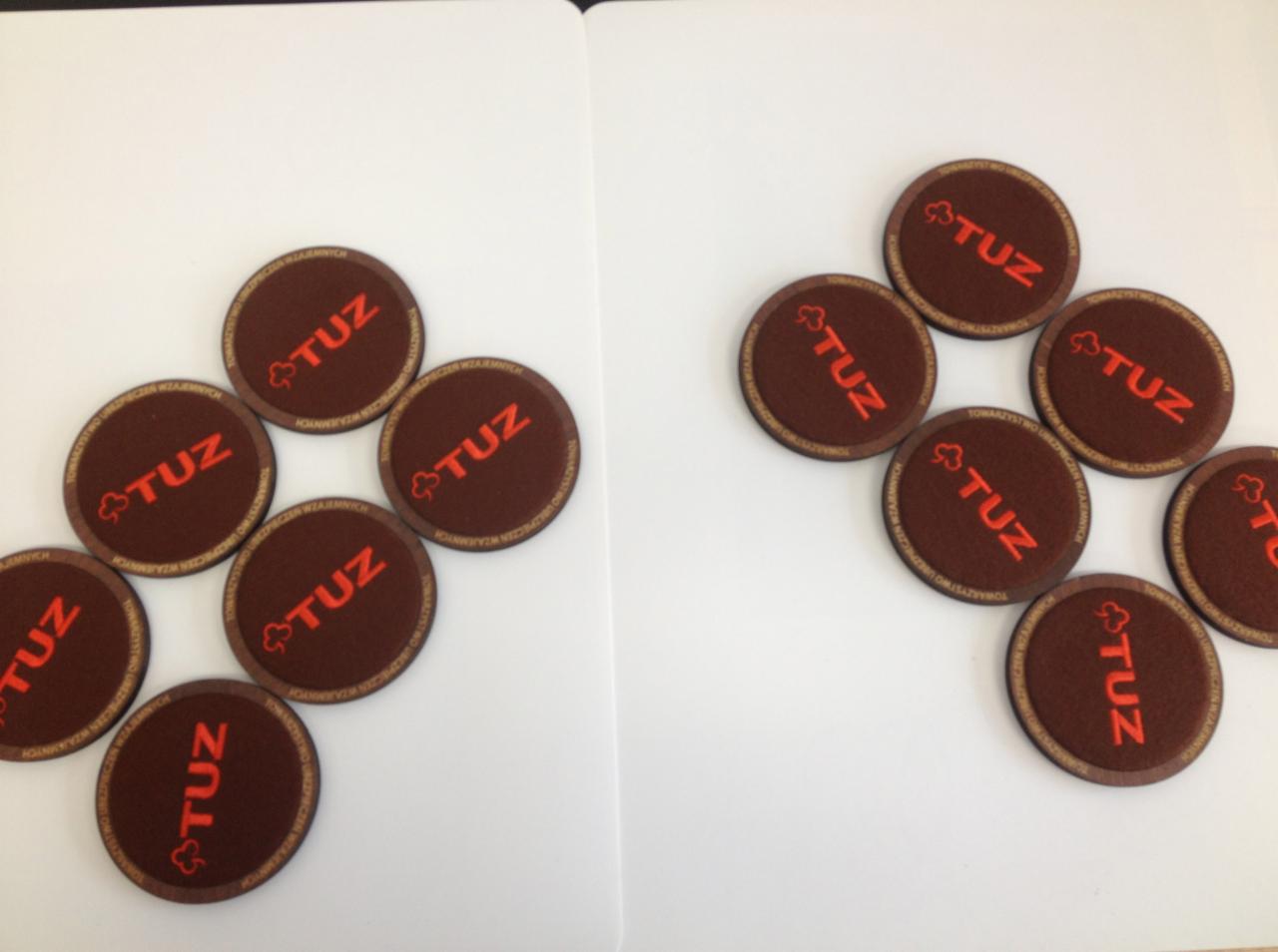 Brązowe podkładki z wyhaftowanym czerwonym napisem 'TUZ' i symbolem koniczyny, ułożone w dwóch grupach na białym tle.
