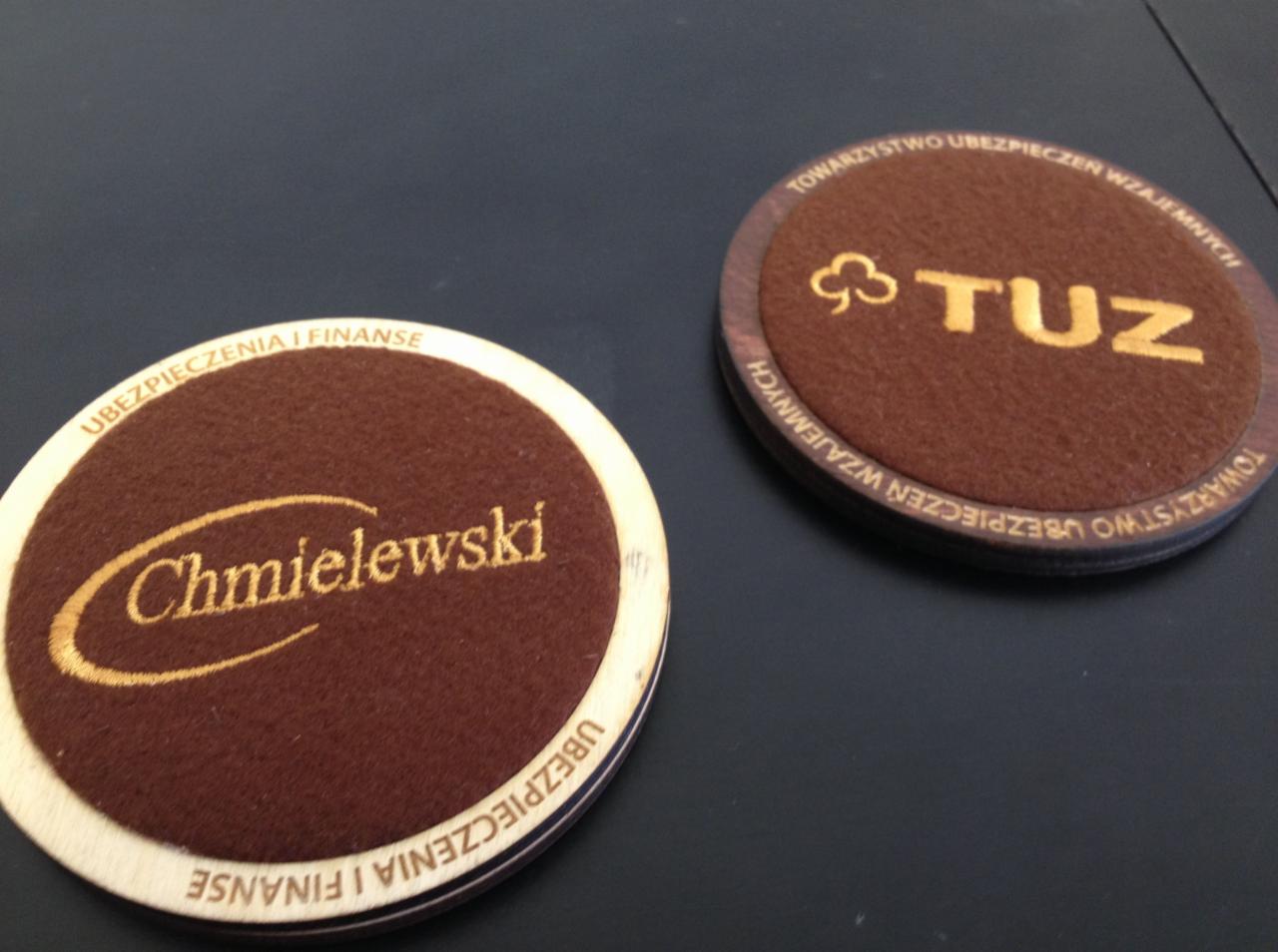 Dwie okrągłe podkładki z brązowego materiału, z wyhaftowanymi złotą nicią napisami 'Chmielewski Ubezpieczenia i Finanse' oraz 'TUZ Towarzystwo Ubezpieczeń Wzajemnych', leżące na ciemnym podłożu.