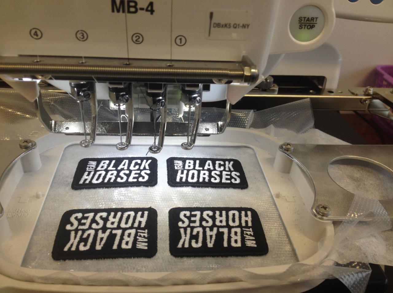Proces haftowania czterech czarnych naszywek z białym napisem TEAM BLACK HORSES, umieszczonych w ramce hafciarki przemysłowej.
