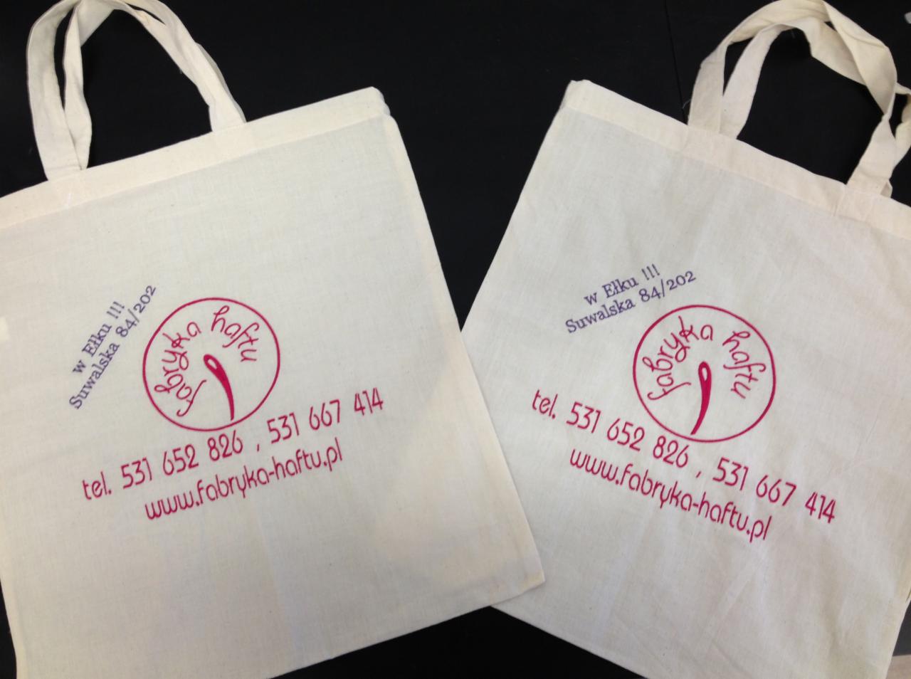 Dwie bawełniane torby z nadrukowanym logo i danymi kontaktowymi firmy Fabryka Haftu w Ełku, widoczne na czarnym tle.