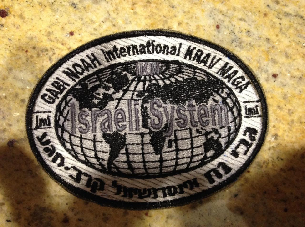 Owalna, haftowana naszywka z motywem mapy świata, napisem 'Israeli System', 'Gabi Noah International KRAV MAGA' i tekstem w języku hebrajskim, leżąca na beżowym tle.