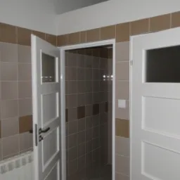 Wykończona łazienka z beżowo-brązowymi kwadratowymi płytkami, białe drzwi z szybą i grzejnik żeberkowy pod ścianą.