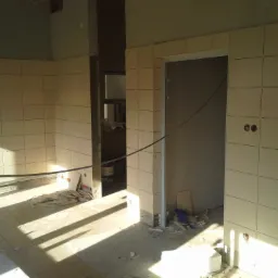 Wnętrze łazienki w trakcie prac: ułożone jasne płytki na ścianach i podłodze, widoczne puste otwory na gniazdka elektryczne, resztki materiałów budowlanych na podłodze i wiadro z klejem w tle.