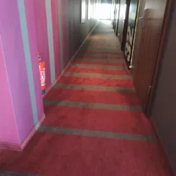 Długi korytarz hotelowy z czerwoną wykładziną w szare pasy. Na ścianie gaśnica. Widok w głąb korytarza, drzwi z numerami pokoi po prawej.