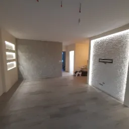 Wnętrze po remoncie: szare panele podłogowe, ściana z dekoracyjną strukturą i podświetleniem LED, wnęki ścienne z oświetleniem, ściana z efektem dekoracyjnym. Widoczne ślady po pracach...