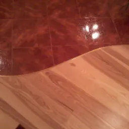 Połączenie brązowych płytek podłogowych i jasnych paneli drewnianych, oddzielone złotą listwą wygiętą w łuk.