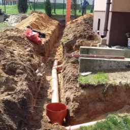 Wykop z ułożonymi rurami kanalizacyjnymi z pomarańczowego PCV, widoczne elementy złączek i studzienki. Wykop w ziemi, w tle budynek i osoba.