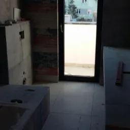 Łazienka w trakcie remontu: szare płytki na podłodze i ścianach, częściowo wykończona zabudowa wanny i blatu, okno balkonowe. Widoczne narzędzia i materiały budowlane.