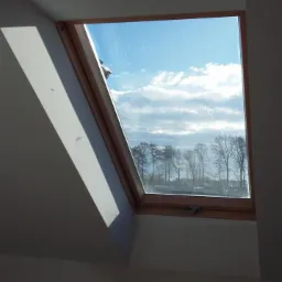 Drewniane okno dachowe z widokiem na błękitne niebo i drzewa. Świeżo pomalowane ściany w odcieniach szarości i bieli. Widoczne promienie słoneczne.