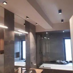 Nowoczesna łazienka z marmurowymi płytkami, punktowym oświetleniem sufitowym i podświetlanym lustrem, widok na kabinę prysznicową za szklaną ścianą.