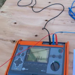 Miernik Sonel MRU-21 z podłączonymi przewodami pomiarowymi, wyświetlający wartość rezystancji uziemienia 39.1 Ω, umieszczony na drewnianej płycie.