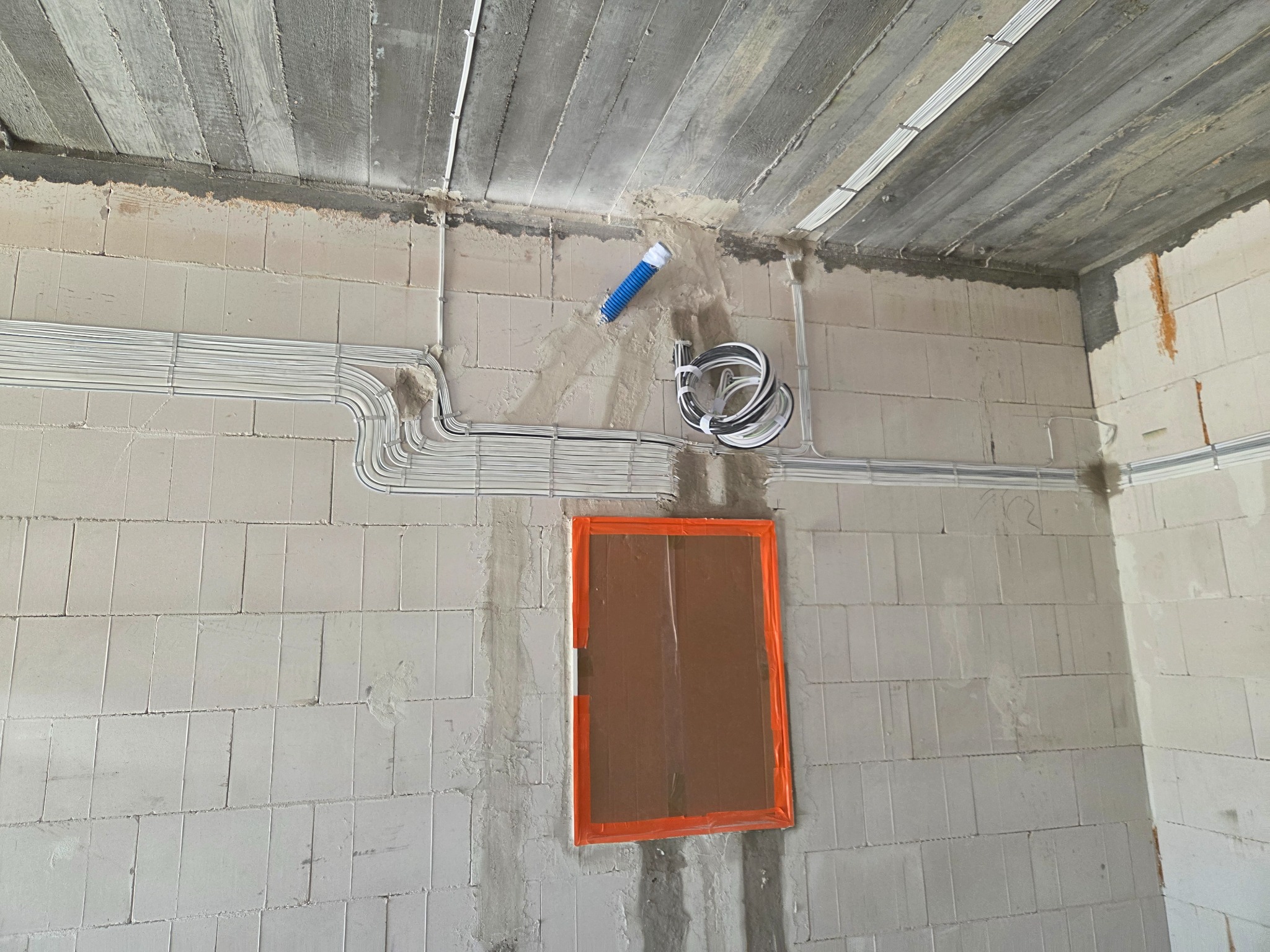 Szare kable elektryczne ułożone wzdłuż surowej ściany z pustaków, z niebieską rurką karbowaną i zabezpieczonym otworem na puszkę elektryczną.