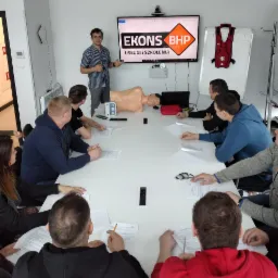 Instruktor w koszuli w paski prezentuje materiały szkoleniowe BHP na ekranie, obok leży manekin do ćwiczeń pierwszej pomocy, a uczestnicy kursu siedzą przy stole z dokumentami.