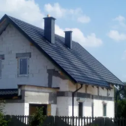 Surowy stan domu jednorodzinnego z białych bloczków, z ciemną dachówką i dwoma kominami, widoczne okna i częściowo otynkowane ściany, ogrodzenie w tle.