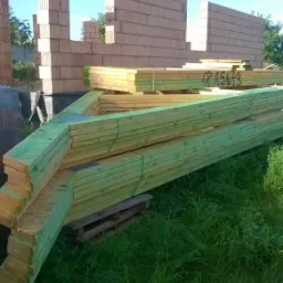 Wznoszone ściany z pustaków ceramicznych na budowie domu, z przygotowanymi elementami drewnianymi na więźbę dachową składowanymi obok.