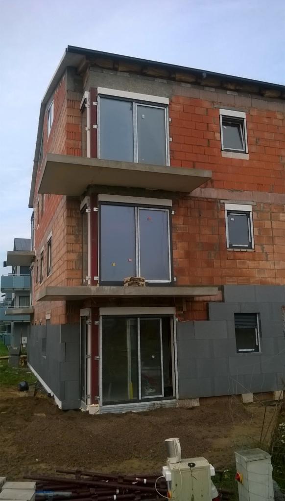 Trzypiętrowy dom w trakcie budowy z czerwonej cegły i szarych bloczków, z zamontowanymi oknami i betonowymi balkonami, widoczne rusztowania i materiały budowlane.