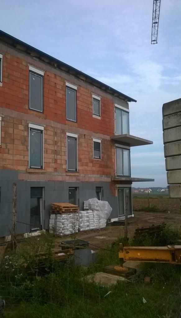 Wznoszony dwukondygnacyjny budynek z czerwonej cegły z szarym ociepleniem na parterze, widoczne okna z roletami, wystające balkony, palety z materiałami budowlanymi i żuraw w tle.