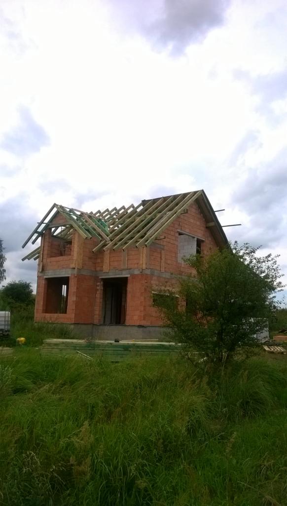 Niedokończony budynek z czerwonej cegły z drewnianą konstrukcją dachu na tle pochmurnego nieba i zarośniętego terenu.