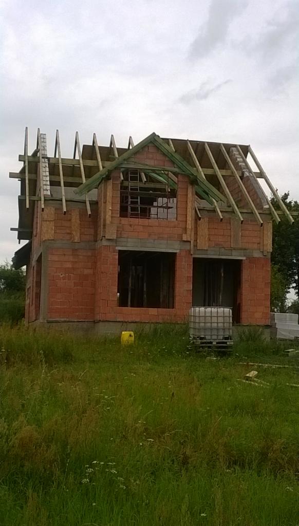 Dwukondygnacyjny dom murowany w stanie surowym otwartym z widocznym szkieletem dachu, zlokalizowany na zarośniętej działce, z pojemnikiem IBC na wodę w pobliżu.